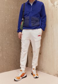 Blå fleecetröja med kvartdragkedja och marinblå fickor, vita cargobyxor med röda detaljer och orangea/vita sportskor.