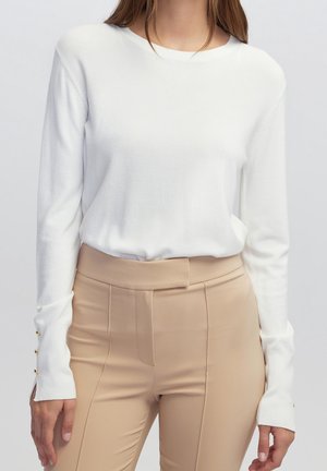 Femme portant un haut blanc uni à manches longues avec des boutons dorés aux poignets et un pantalon beige taille haute sur un fond uni.