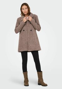 Brauner strukturierter Doppelreiher-Mantel mit Kragen, schwarzen Knöpfen und einer lockeren Passform, kombiniert mit engen schwarzen Hosen und braunen Stiefeletten.