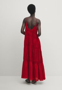 Punainen, hihaton maksimekko, jossa on syvä V-muoto takana, ja siinä on kerrostettu helma sekä kerääntynyt pinta. Materiaali: kevyt kangas.