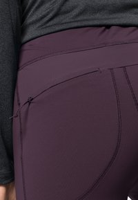 Lila högmidjade leggings med en slät yta, utrustade med en sidficka och kontrasterande sydda sömmar. Kombineras med en grå långärmad tröja.