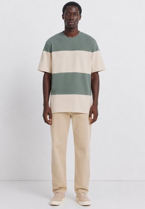 Junger Mann stehend, trägt beige Hosen, beige-grün breit gestreiftes kurzärmeliges Hemd und beige Schuhe vor einem einfarbigen weißen Hintergrund.