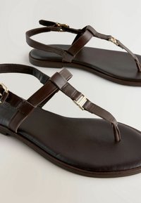 Sandales plates en cuir marron avec brides autour de la cheville et détails de boucles dorées, conçues pour un usage décontracté.