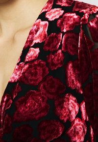 Tessuto nero con un motivo floreale in texture di velluto rosso. Il design presenta grandi rose vivide con petali lisci su uno sfondo scuro.