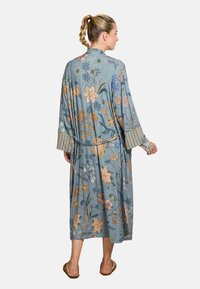 Woman with braided hair wearing a long, blue floral robe with striped cuffs and slippers, standing facing away against a plain background.