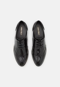 Chaussures noires en cuir verni à lacets avec des détails de brogue, présentant un bout arrondi et une finition lisse et brillante. L'intérieur affiche le nom de la marque.