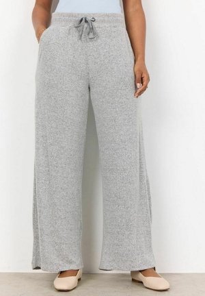 Femme portant un pantalon large gris clair avec une taille à cordon et des chaussures plates beiges, debout contre un mur blanc.