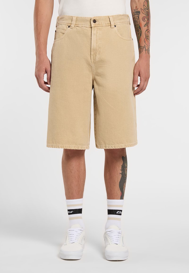 Pantalones cortos de mezclilla beige con corte recto, cinco bolsillos y costuras visibles. Usados con zapatillas blancas y calcetines a rayas.