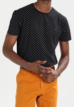 Schwarzes T-Shirt mit kurzen Ärmeln und weißen Punkten, runder Ausschnitt und einem kleinen blauen Logo auf der Brust, getragen mit orangefarbener Hose.