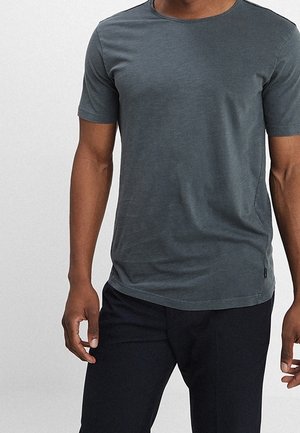 Homme portant un t-shirt gris uni à manches courtes et un pantalon noir, debout devant un fond clair.