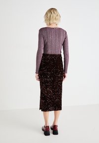 Haut violet à manches longues, côtelé, associé à une jupe marron à sequins arrivant au genou. Le mannequin porte des chaussures noires à talons épais. Fond uni.