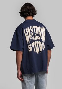 Marineblauw oversized katoenen T-shirt met grote crèmekleurige grafische tekst "WRSTBHVR STUDIO" op de achterkant, met een relaxte pasvorm en korte mouwen.