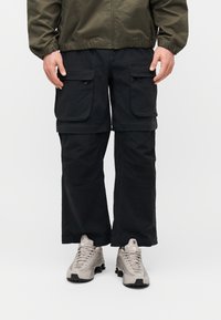 Κάτω μέρος σώματος ενός ατόμου που φοράει μαύρο παντελόνι cargo με μεγάλες τσέπες και ανοιχτογκρι αθλητικά παπούτσια σε απλό λευκό φόντο.