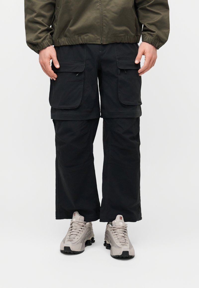 Κάτω μέρος σώματος ενός ατόμου που φοράει μαύρο παντελόνι cargo με μεγάλες τσέπες και ανοιχτογκρι αθλητικά παπούτσια σε απλό λευκό φόντο.