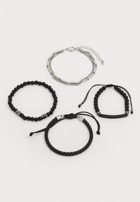Cuatro pulseras: una cadena de plata, una pulsera de cuentas negras con acentos metálicos, un estilo trenzado de cuero negro, y una banda delgada negra con cuentas.