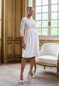Žena v bielych šatách po kolená s čipkovými rukávmi a v sandáloch na vysokom podpätku stojaca vedľa ozdobného krémového kresla pri okne.