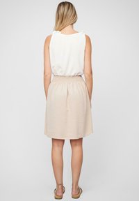 Haut sans manches blanc avec des accents à nouer aux épaules, associé à une jupe en tissu léger de longueur genou, taille smockée, de couleur beige.