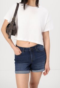 Vit kort t-shirt kombinerad med blå denimshorts med uppvikta fållar. Accessoarer inkluderar en liten brun läderväska som bärs tvärs över kroppen.