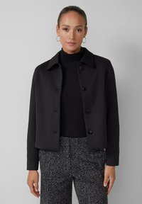 Schwarze, kurze Jacke mit Kragen, vorderem Knopfverschluss und strukturiertem Stoff, getragen über einem gerippten Rollkragenpullover.