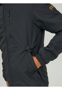 Czarna kurtka z zapinaną na zamek kieszenią na piersi, elastycznymi mankietami i rękami w bocznych kieszeniach; mała pomarańczowa metka marki na górnej części rękawa.