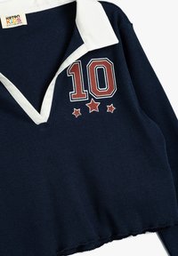 Marineblå ribbet langærmet polo med hvid krave, prydet med et broderet rødt tal "10" og tre stjerner nedenfor. Beskåret kantdetalje.