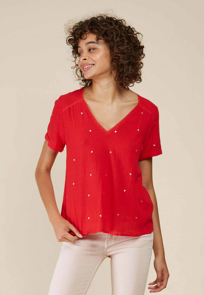 Blouse rouge à manches courtes avec un col en V, décorée de petits motifs triangulaires blancs. Tissu léger avec une texture douce.