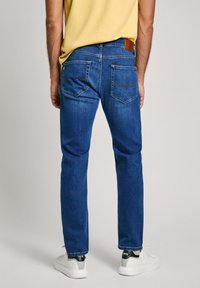 Jeans en denim bleu avec une coupe slim, présentant une patch en cuir sur la taille et quatre poches, portés avec des baskets blanches.