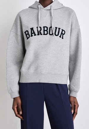 Personne portant un sweat à capuche gris clair avec un texte "BARBOUR" en noir et un pantalon bleu foncé, debout devant un fond clair uni.