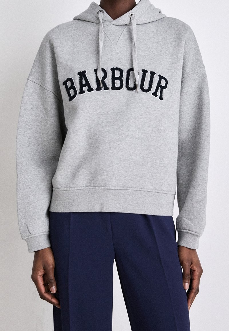 Personne portant un sweat à capuche gris clair avec un texte "BARBOUR" en noir et un pantalon bleu foncé, debout devant un fond clair uni.