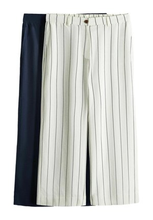 Kobieta stojąca w luźnej czarnej koszuli na czarnym topie, białe spodnie o szerokich nogawkach z pionowymi czarnymi paskami, czarny pasek i czarne buty na jednolitym tle.