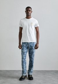 Λευκό μπλουζάκι κοντό μανίκι με στρογγυλή λαιμόκοψη και κυκλικό λογότυπο. Μπλε τζιν με λευκά σχέδια τύπου γκραφίτι. Μαύρα αθλητικά παπούτσια.