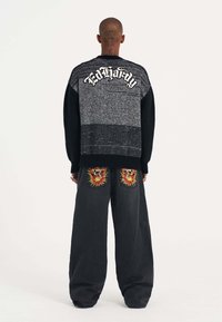 Černý a šedý pletený svetr s logem "Ed Hardy" na zádech, spárovaný s volnými džíny zdobenými barevným výšivkami lebek a slunečními paprsky.