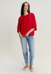 Rode oversized top met raglanmouwen, gecombineerd met lichtblauwe jeans. De outfit wordt aangevuld met beige platte schoenen.