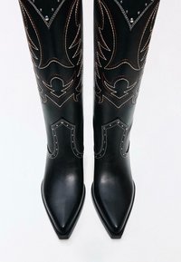 Zwarte leren kniehoge laarzen met puntige neuzen, voorzien van ingewikkeld stikwerk en zilveren accenten. Het ontwerp bevat gedurfde, decoratieve patronen.