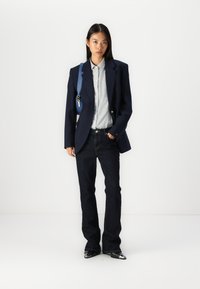 Blazer marine, chemise rayée bleu clair, jeans en denim noir, chaussures noires brillantes. Le blazer présente des boutons dorés et un design structuré.