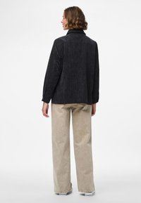 Žena s kratkom valovitom kosom, nosi crnu kordiranu košulju i bež široke hlače, stoji okrenuta leđima prema običnoj bijeloj pozadini.