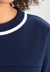 Mörkblå sweatshirt med en vit ribbad krage och en slät yta. Har en något upphöjd söm över br östområdet.