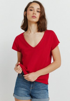 Młoda kobieta o brązowych włosach do ramion, ubrana w czerwoną koszulkę z dekoltem w serek i podwinięte niebieskie szorty dżinsowe, trzymająca brzeg swojej koszulki.