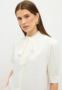 Blusa bianca con fiocco al collo, maniche corte a sbuffo e chiusura frontale con bottoni. Tessuto morbido con una leggera lucentezza. Orecchini a cerchio dorati.