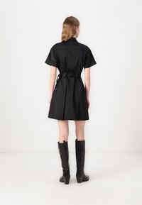 Vestuário preto de mangas curtas com uma cintura cingida, apresentando um corte reto e bolsos laterais. A modelo usa botas brilhantes pretas até ao joelho.