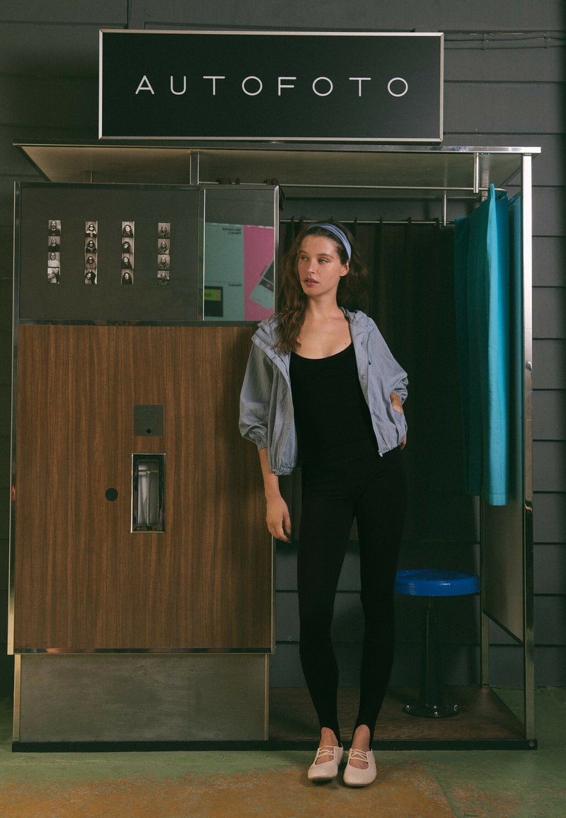 Jeune femme portant une tenue noire et une veste légère, debout à côté d'une cabine photo vintage avec un rideau et un tabouret bleu à l'intérieur.