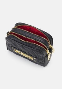 Proš�ívaná černá taška z umělé kůže se zlatým kováním, dvěma kapsami na zip a červenou vzorovanou podšívkou s logem "LOVE MOSCHINO".