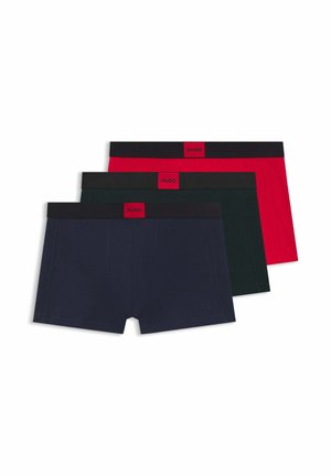 Młody mężczyzna z krótkimi ciemnymi włosami, ubrany w granatowe bokserki z czarną gumką i czerwoną metką "Hugo", stojący na jednolitym białym tle.