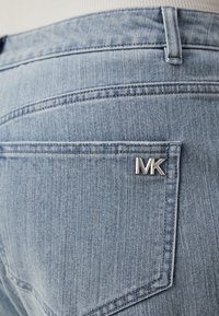 Vaaleansiniset farkut esittelevät takataskua, jossa on hopeinen MK-logo. Kankaassa on hienovarainen tekstuuri ja tasainen ompeluyksityiskohta.