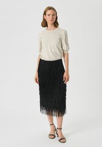 Czarna spódnica z frędzlami o teksturowanej powierzchni, połączona z jasnobeżową bluzką z krótkim rękawem. Uzupełniona czarnymi sandałami na obcasie.