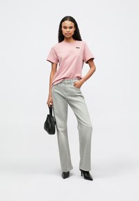 Διαθέτει ροζ κοντομάνικο μπλουζάκι με μικρό λογότυπο, συνδυασμένο με ψηλόμεσα ανοιχτό γκρι φαρδιά παντελόνια και μαύρα τακούνια, κρατώντας μια μαύρη τσάντα.