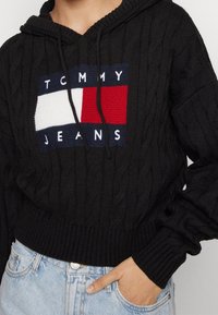Black cable-knit hoodie with Tommy Jeans logo in white, navy, and red, worn with light blue denim jeans, hand placed in pocket.