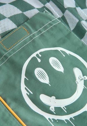 Grüner Stoff mit einem großen weißen Smiley-Face-Graphic, Skateboard-Details und orangefarbenen Stitching-Akzenten, vor einem karierten Mesh-Hintergrund.