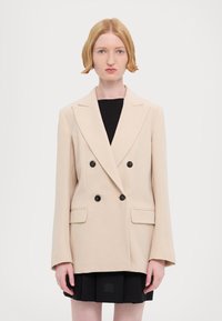 Beige bleiseri kaksiriviseen nappiin mustilla napeilla, jossa on kauluskanta, kaksi etutaskua ja rento leikkaus. Vaatteena mustan mekon päällä.