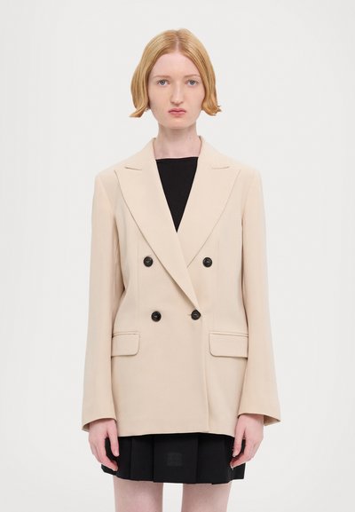 Blazer beige double-breasted avec des boutons noirs, doté d'un col cranté, de deux poches avant et d'une coupe décontractée. Porté par-dessus une robe noire.